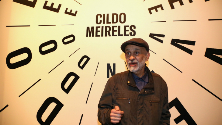 O artista Cildo Meireles posa em frente ao painel da sua exposição. Crédito: Denise Andrade